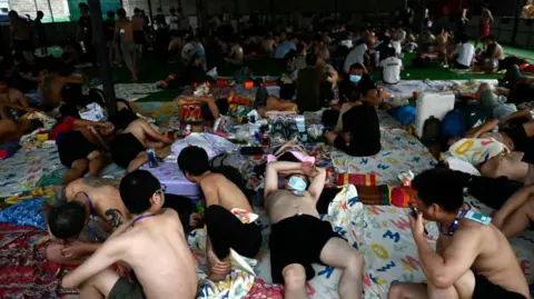 Getty pictures of a group of men, some of whom are lying on Mattresses, while others sit or stand, in a temporary camp. Many of them without a shirt and in short pants. 