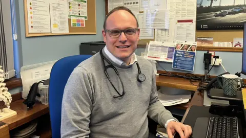 Isabella Verona/ BBC Jamie Green is seated at a desk in what appears to be a medical office or consultation room. Green is wearing a light grey sweater over a collared shirt, and a stethoscope hangs around the neck. The desk holds a computer keyboard, a telephone, paperwork, and a patterned mug. Behind Green, a wall-mounted board is filled with printed charts, notices, and medical information. To the left, anatomical models and binders are visible on a wooden shelf. The room has a clinical, organised appearance, with various documents and medical materials displayed throughout the workspace.
