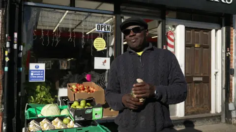 Wayne Lawrence wearing a flat cap and sunglasses, in dark clothing, smiling outside Progress Barbers, standing next to stables stacked with groceries.