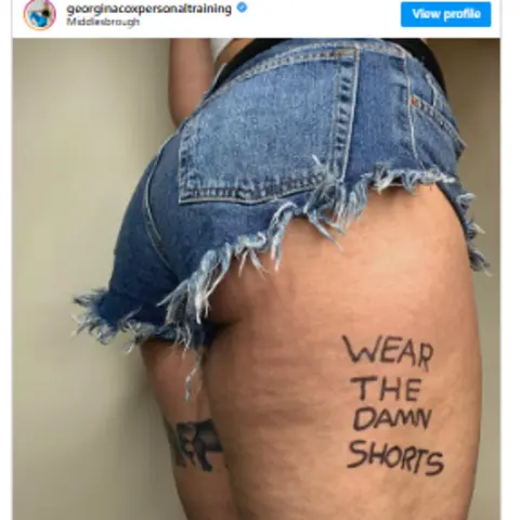 Georgina Cox Instagram post by Georgina Cox, showing her body from the waist to mid thigh. She's wearing a pair of very short cut-off jeans shorts, with a frayed hem. A little of her bottom is showing and on her average-sized and strong-looking upper thigh are the words "Wear the damn shorts".