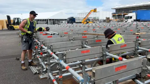 Three construction workers working on structures at the Farnborough International Exhibition & Conference Centre ahead of this year's event. They are wearing hats and high vis jackets. Operational machines and lorries are parked nearby.
