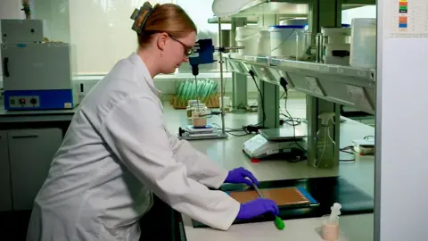 A woman in a white lab coat and purple gloves is leaning over a counter in a lab. She is using a metal bar to spread a thick, beige coloured solution over a piece of brown card. To the side is a glass beaker full of the solution with a syringe standing in it. The scientist has her long blonde hair tied back in a clip and is wearing goggles over her glasses. 