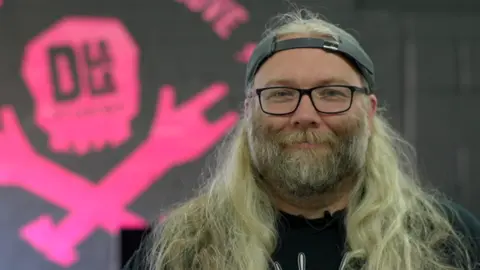 Jamie Niblock/BBC AJ Grand-Scrutton has long blond hair and is wearing a cap backwards. He also has glasses on. There is a pink logo on the wall behind him.