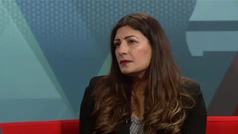 Preet Kaur Gill wears a black jacket with a black and white garment underneath. She looks to the left of the camera with a solemn expression. She sits on a red sofa with a blue backdrop behind her.