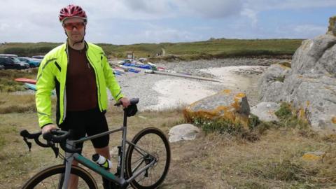 Josh Paul has a hi-vis coat, is wearing a cycle helmet and is holding his bike in what looks like a quarry, with a cycle path in the background