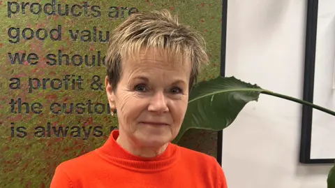 Helen Ambrosen has dark hair with blonde highlights in it and is looking at the camera and smiling. She is wearing a bright orange jumper and in front of what appears to be Lush's mission statement. 