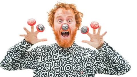 A man with red hair and beard is dressed in a black and white top, featuring cartoon drawings of a variety of characters. He is wearing one red nose with black and white doodle illustrations, and four of his fingers with a red nose with black and white doodle illustrations.