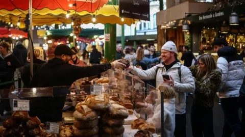 Getty Images A bread stall in a crowded market. A baker hands over a loaf to a customer.