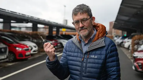 BBC A man with dark hair, a light beard wearing a blue jacket, standing in front of parked cars with a car key in his hand