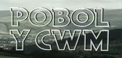 'Pobol y Cwm' in white font is displayed in front of an image of a green valley