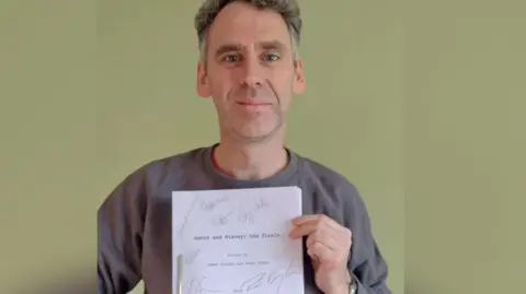 Robert Wilfort, who plays Jason in Gavin and Stacey, smiles at the camera holding the Christmas Day episode finale script which has been signed bby all the main cast. Robert has grey hair and is wearing a grey jumper. 