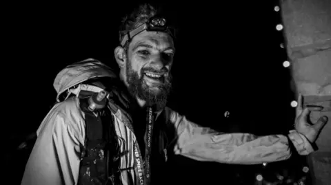 Wild Aperture A picture with a black-and-white filter showing runner Rhys Beddoe at the finish line of the Spine Race. He has a beard and is wearing a headtorch, a backpack and a waterproof.