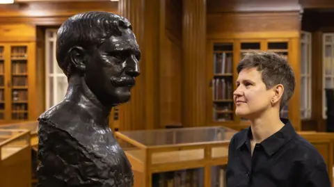University of Leeds A person with short brown hair wearing a black shirt looks at a bronze bust statue of a man with a moustache and short hair. They are in a library.