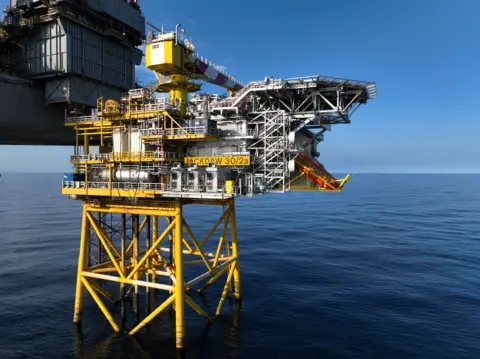 A mostly yellow-painted gas platform and allied grey rig stand in calm blue seas with a clear blue sky behind them. It is sunny.