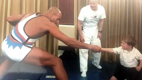 Daz Crawford Crawford, wearing his white, black, red and blue gladiator singlet is taking part in a hand-holding tug of war with a young boy. Behind them is a martial arts instructor.