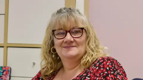 Gatehouse A head and shoulders image of a woman with shoulder length blonde hair. She is wearing gold, hoop earrings, glasses and a red, white and black floral top. She is looking directly into the camera. 