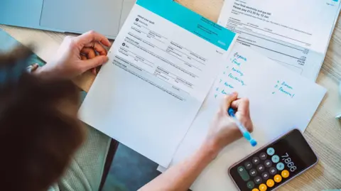 Getty Images Overhead shot of woman looking at energy bills with pen in hand and calculator nearby.