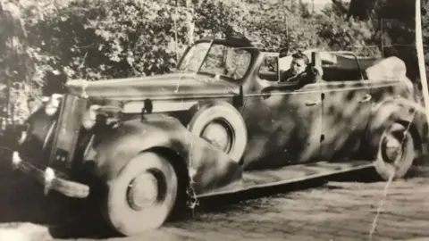 John Lloyd A black and white photo from spring 1945. It shows Jack Lloyd sitting behind the wheel of an open top German officers' car. Behind it is a shrubbery. 