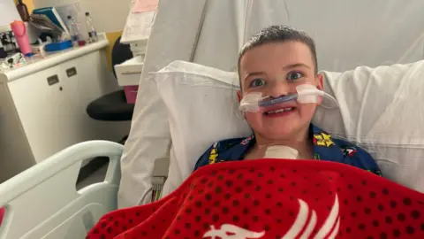 Máirtín Mac Gabhann A young boy is pictured in a hospital bed. He is dressed in pyjamas and has a red patterned blanket on top. He has 