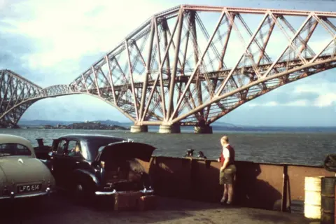 Ray Stokes Man next to old car with its boot open with large iron bridge over water in the background