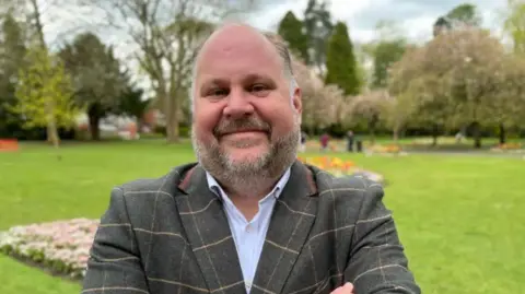 Leader of Swindon Borough Council, Jim Robbins, stood in what looks like a park. There are flower beds, tress and bushes behind him. He smiling and folding his arms in front of his chest. 