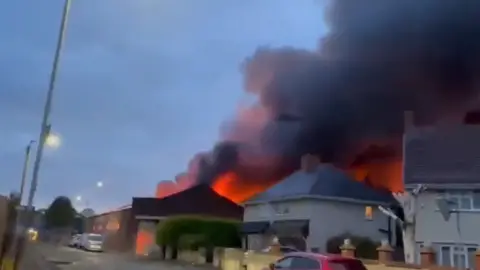 A large fire can be seen behind a street of semi-detached houses. Smoke and flames are rising above the height of the rooftops. There are parked cars on the road in front of the houses.