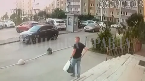 A man in jeans and a dark t-shirt holding a plastic bag and a holdall in one hand walks out of a building towards a car park in Kyiv.