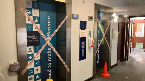 Two lift entrances sealed with tape across them. The tape says "Contents checked and security sealed". Orange and yellow cones have also been placed in front of them