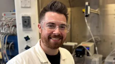 Josh Bailey is a young man with a short reddish/brown bears and brown hair which is shorter on the sides and longer on top. He is wearing a white lab coat over a black t-shirt and a pair of plastic safety goggles. He is standing in front of some lab equipment and is smiling at the camera.