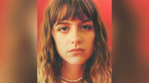 Brits Fringe handout Chloe Slater with long brown hair wearing a silver nose ring and pearl necklace and cream top. She is pictured in front of a red background and looking impassively at the lens.