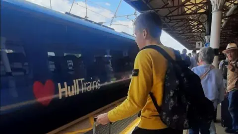 Olesia Khrapova Maksym stands on a platform at Hull train station and is wearing black shorts and a yellow top. He has a blue suitcase and a black rucksack on his back. 