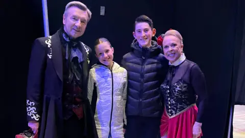 The two cousins pose for a picture with Torvill and Dean. Isla is in a white quilted jacket, her light brown hair in a ballet bun, Lewis has a smart short hairstyle and a black puffer jacket, while Torvill and Dean stand on the outside, wearing skating costumes in black, red and gold and in a Baroque style.
