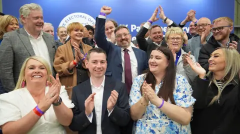 PA Media Robin Swann with an arm held aloft stands in the middle of happy, clapping supporters 