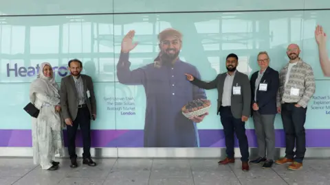 Heathrow Airport Ltd Shah stands to the right of his Heathrow poster with two other men next to him. On the left are his parents. The poster shows Shah holding a bowl of dates in one hand and waving with the other
