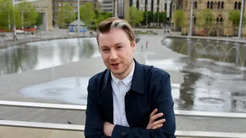 Si Cunningham - a man in a blue suit jacket and white shirt stands in front of Bradford's city park. He has brown hair with a white streak in the front