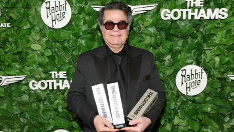 Getty Images Jafar Panahi in a black suit and dark glasses holding three awards saying "The Gothams", in front of an ivy-covered backdrop with various logos