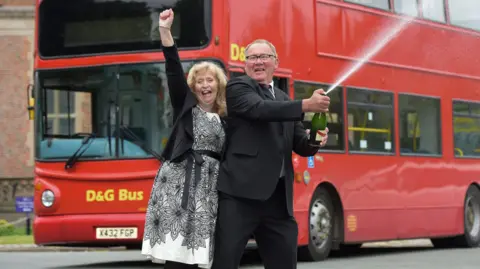 A man and a woman are celebrating in front of a red double-decker bus. The man is spraying champagne from a bottle, while the woman has her arm raised in the air. 