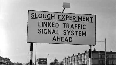 A black and white photo shows a large road sign with the words 'Slough Experiment, linked traffic signal system ahead' written in black on a white background. Behind the sign are the top of a row of terraced buildings and the top of a double decker bus can be seen to the side of them.