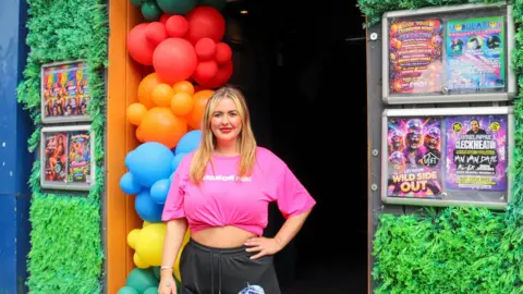 BBC / JOSH PARRY A woman stands in the doorway of a venue decorated with a large rainbow balloon arch, wearing a bright pink cropped T‑shirt and black shorts. The entrance is framed by green artificial foliage walls and notice boards filled with colourful event posters, including listings for club nights and performances at The Loft in Cleckheaton. The street setting is just visible at the edges, while the dark interior of the venue contrasts with the vibrant decorations at the entrance