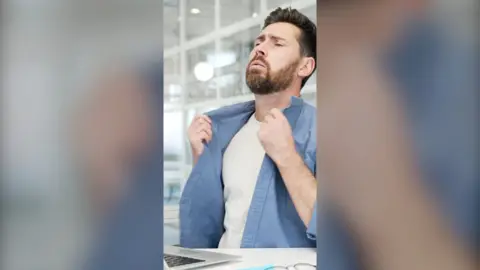A man holds his shirt collar and looks uncomfortable at a desk