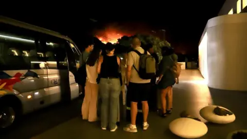 EPA Tourists stand around next to a minibus outside a hotel after evacuating during a wildfire in Ierapetra, on Crete island