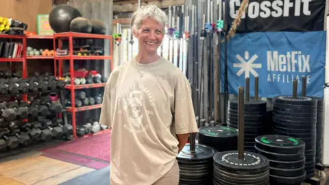 Krish Stevens A woman with grey hair smiling and standing in front of gym equipment including free weights