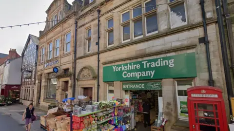 Google Stone buildings on a shopping street. The closest has a green sign over the door saying Yorkshire Trading Company. Outside it is a red public telephone box and stands stacked with goods including plant pots. Next door is a building with a blue Barclays sign above the window. 