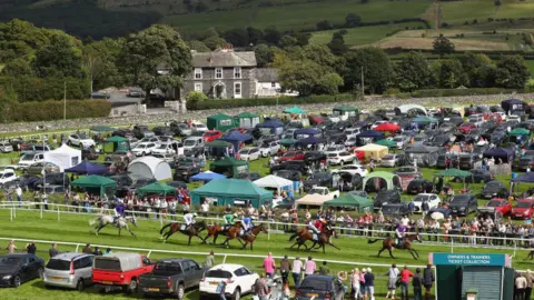 Getty Images Arena Balap Cartmel digambarkan selama perlombaan Handicap Hurdle Hadwins Motor Group pada tahun 2017. Kuda sedang berlomba dan lapangan dikelilingi oleh mobil dan gazebo.