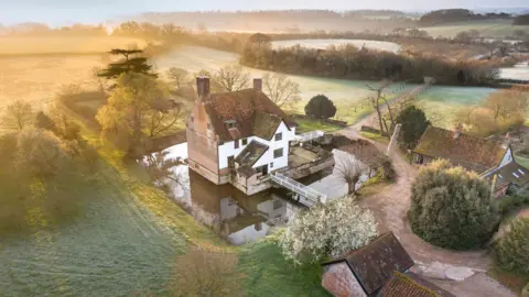 Historic England Archive An aerial image of Letheringham Lodge, showing the former hunting lodge and moat. It is surrounded by vast plots of empty land as well as other, smaller buildings and trees. 