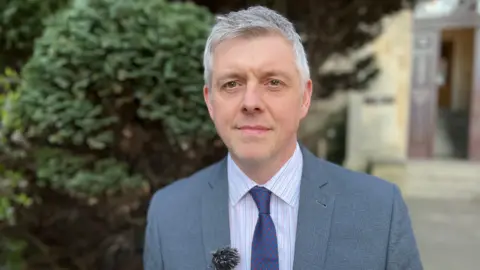 A man with short grey hair stands outside a court. He is wearing a grey suit, a striped shirt and a blue tie which has red spots on it, along with a lapel microphone.