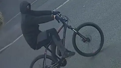West Midlands Police A man on a black bicycle who is dressed completely in black, including gloves and a balaclava