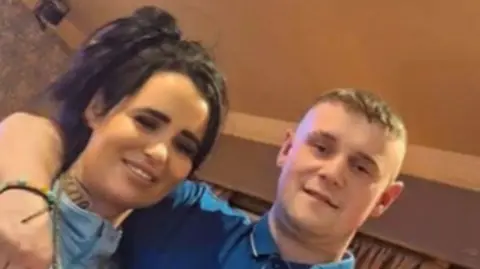 A close-up grainy photo of Nathan Bushby and his sister. Nathan has short light brown hair and is smiling. He is wearing a blue polo top and has his arm around his sister who has long black hair and is wearing a light blue top. 