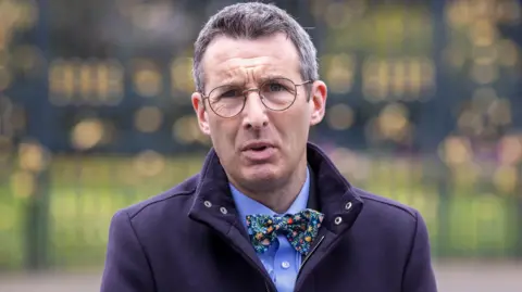 PA Media Andrew Muir, he is wearing glasses with a navy coat, blue shirt and floral bowtie. He has dark grey hair.