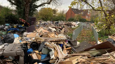 PA Media A huge mountain of waste including lots of bits of wood, car seat, sofa on its side and mattresses on a grass field in front of some houses